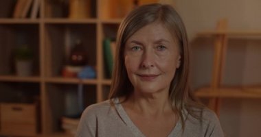 Kameraya bakan yaşlı, çekici, sarı saçlı kadının yakın görüntüsü. Yaşlı kadın evde otururken poz veriyor. İnsanların vesikalık fotoğrafı, rahatlama ve rahatlama kavramları.