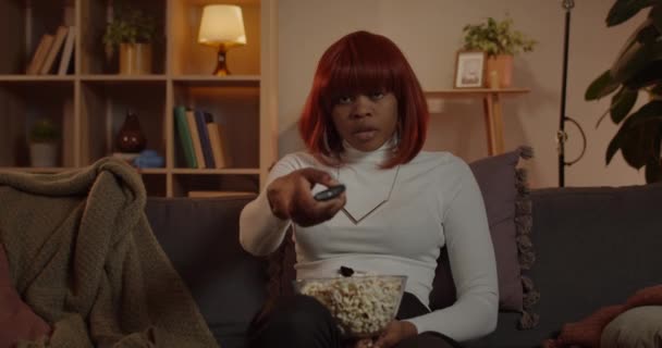 Vista de la cosecha de la hermosa mujer que toma el mando a distancia y encender la televisión. Mujer afroamericana sosteniendo tazón con palomitas de maíz mientras está sentada cómodamente en el sofá y viendo películas. — Vídeo de stock