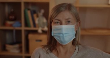 Tıbbi koruyucu maskeli yaşlı bir kadının portresi başını çevirip kameraya bakıyor. Sarışın bir kadının evde oturduğu yakın plan görüntüsü. Karantina ve virüs kavramı.