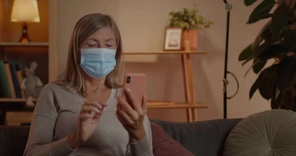 Crop view of elederly woman in medical protective mask touching smartphone screen while sitting on sofa at home.Female mature person with short blond hair while using phone. — Stock Video