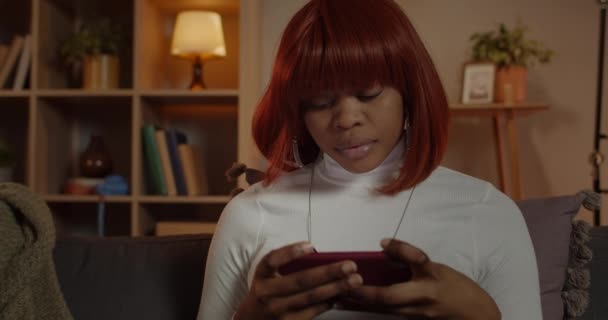 Vista de la cosecha de la mujer joven viendo vídeo mientras está sentado en el sofá en casa. Persona femenina afroamericana con cabello rojo sosteniendo el teléfono inteligente horizontalmente mientras mira la pantalla. — Vídeos de Stock