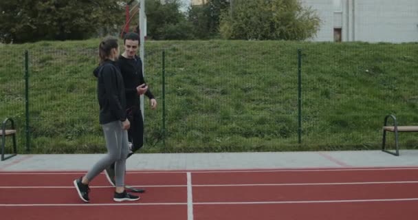 一个带着假肢跑步刀片的年轻人在室外和女人说话的侧视图。腿被截肢的男性与女运动员聊天，在运动场上散步时微笑. — 图库视频影像