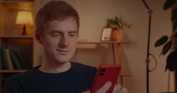 Close up of joyful red haired man scrolling screen and checking phone while sitting on sofa at home. Handsome younng guy looking surprised and rejoicing while doing yes gesture. — Stock Video