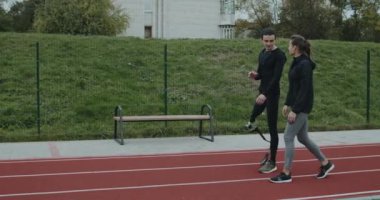 Protez koşu bıçaklı neşeli bir adamın açık havada kadınlarla konuşmasının yan görüntüsü. Ampüte edilmiş bacağı olan erkek sporcu kadın oyuncuyla sohbet ediyor ve spor sahasında yürürken gülümsüyor..