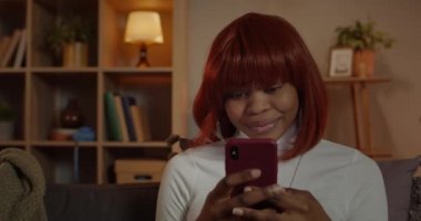 Evdeki koltukta otururken akıllı telefon kullanan ve gülümseyen güzel bir kadın görüntüsü. Afro Amerikalı kadın sosyal medyada haber yayını yapıyor ve telefon ekranına bakıyor..