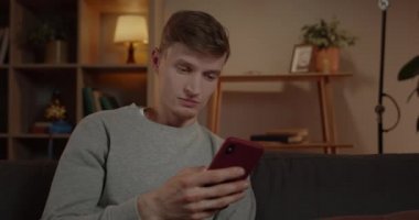 Evdeki kanepede otururken milenyum tırnağı adamın telefon ekranını kaydırmasının ekili görüntüsü. Yakışıklı erkek şaşırmış görünüyor ve akıllı telefonu kullanırken ve kontrol ederken vay canına diyor..