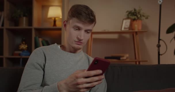 Crop view of amazed millennail man scrolling phone screen while sitting on sofa at home. Bonito homem pessoa olhando surpreso e dizendo uau ao usar e verificar smartphone. — Vídeo de Stock