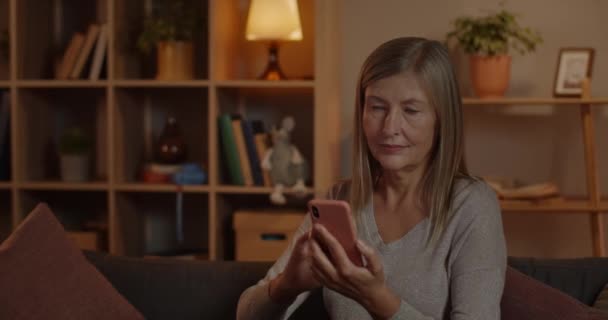 Mujer tocando la pantalla del teléfono inteligente mientras está sentado en el sofá en casa. Persona bastante femenina con pelo rubio corto sonriendo mientras usa y desplaza el teléfono. — Vídeos de Stock