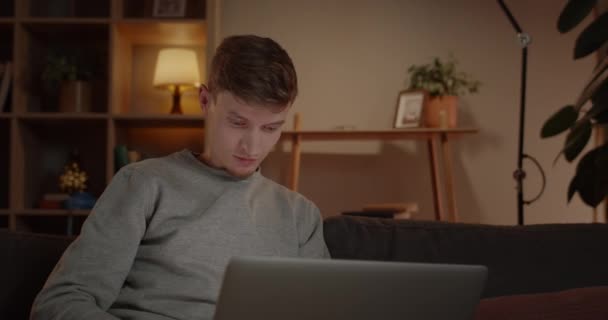 Vista Crop di giovane freelance utilizzando il computer portatile mentre seduto sul divano a casa. Persona maschile concentrata con i capelli ondulati che lavorano online e guardando lo schermo del dispositivo. Concetto di lavoro a distanza. — Video Stock