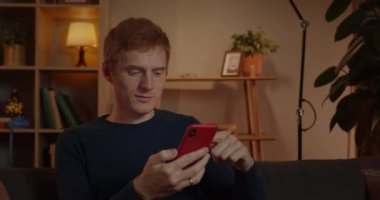 Heyecanlı kızıl saçlı adamın evdeki kanepede otururken ekranda kayan görüntüsü. Akıllı telefonu kullanırken ve kontrol ederken şaşkın ve neşeli görünen neşeli erkek..