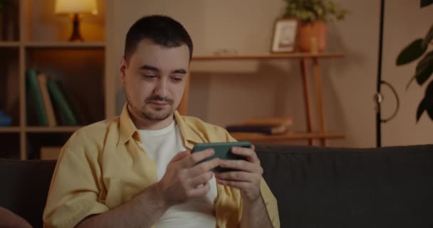 Crop view of concentrated man watching video while sitting on sofa in evening at home. Adult male person holding smartphone horizontally and looking at screen. Concept of leisure. — Stock Video