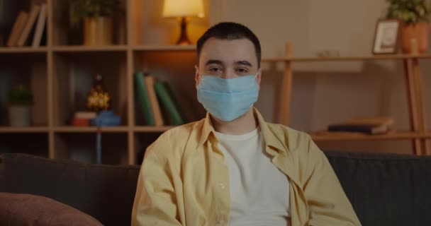 Portret van een man met een medisch beschermend masker die hoofd draait en naar de camera kijkt. Crop view van mannelijke volwassene thuis zitten. Concept quarantaine, virussen, gezondheidspreventie. — Stockvideo