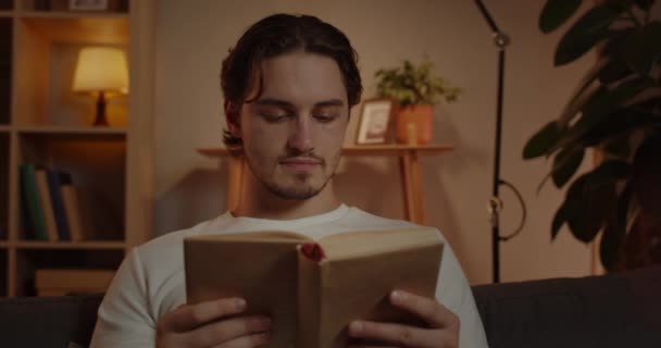 Nahaufnahme einer intelligenten männlichen Person, die liest und ihre Freizeit verbringt. Schöner junger Mann genießt das Buch, während er auf dem Sofa im gemütlichen Heim sitzt. Konzept von Mensch und Freizeit. — Stockvideo
