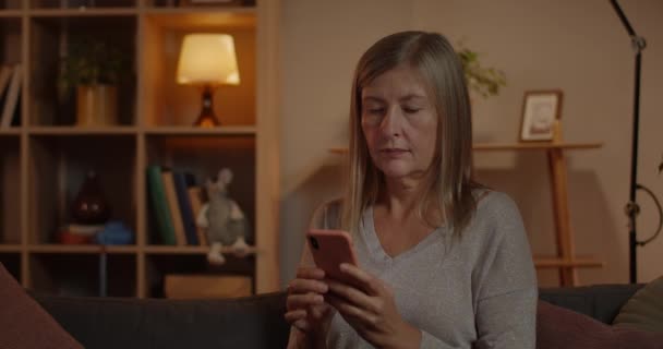 Crop view of serious woman typing message at smartphone screen while sitting on sofa at home. Beautiful elderly woman with short blond hair using phone while spending free time. — Stock Video