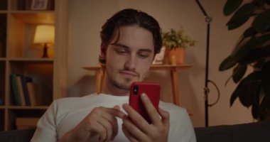 Evdeki kanepede otururken akıllı telefon kullanan yakışıklı bir adamın görüntüsü. Sosyal medya haberlerini tarayan ve telefon ekranına bakan adam. Boş zaman ve boş zaman kavramı..