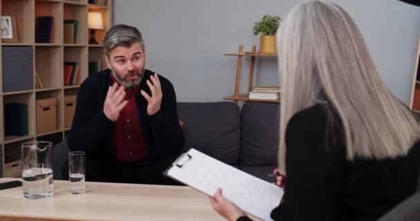 Crop view of emotional man sitting and having therapy session with female person. Homme barbu parler des problèmes et des gestes tandis que la femme thérapeute prendre des notes. — Video