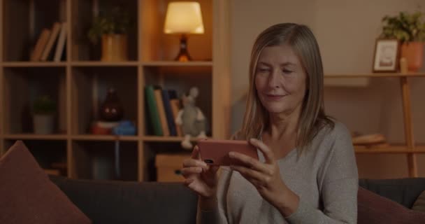 Вид на літню жінку, яка дивиться відео і посміхається, сидячи на дивані вдома. Весела жінка з світлим волоссям тримає смартфон горизонтально, дивлячись на екран . — стокове відео