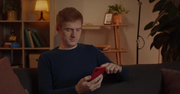 傍晚时分，年轻的红头发男人坐在沙发上滚动智能手机屏幕的照片。一个英俊的男人在业余时间用手机。休闲的概念. — 图库视频影像