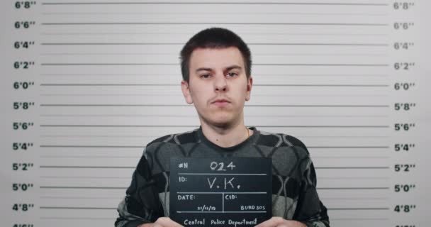 Portrait of male criminal holding sign for photo in police department. Mugshot of young man posing, raising head and looking to camera in front of lineup metric wall. Concept of crime. — Stock Video