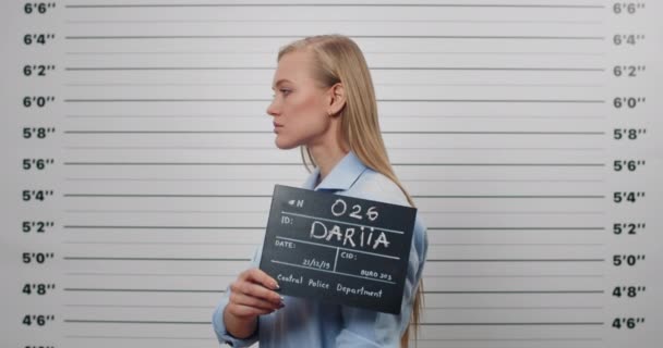 Zdjęcie atrakcyjnej kobiety trzymającej tabliczkę ze zdjęciem na posterunku policji odwracając się na bok. Kobieta przestępca patrząc do kamery, stojąc przed metryczną ścianą składową. — Wideo stockowe