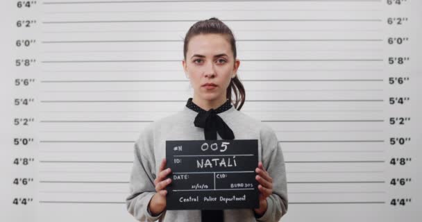 Portrait of young woman with ponytail holding sign for photo in police department. Crop view of pretty female criminal posing, raising head and looking to camera.Concept of mugshot. — Stock Video