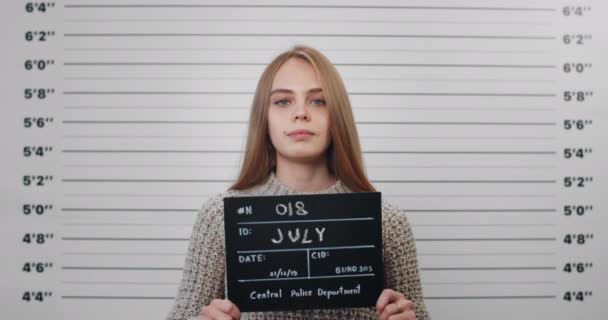 Portrait of young woman with long blond hair holding sign for photo in police department. Crop view of you female criminal posing, raising head and looking to camera.Concept of mugshot. — Stock Video