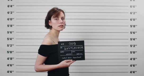Portrait of arrested millennial woman turning head and looking to camera while standing aside. Female person with short haircut holding sign for photo in police department. Concept of mugshot. — Stock Video