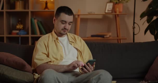 Вид на человека, прокручивающего экран смартфона, сидя на диване в гостиной. Расслабленный мужчина пользуется телефоном, проводя свободное время дома. Концепция досуга и развлечений. — стоковое видео