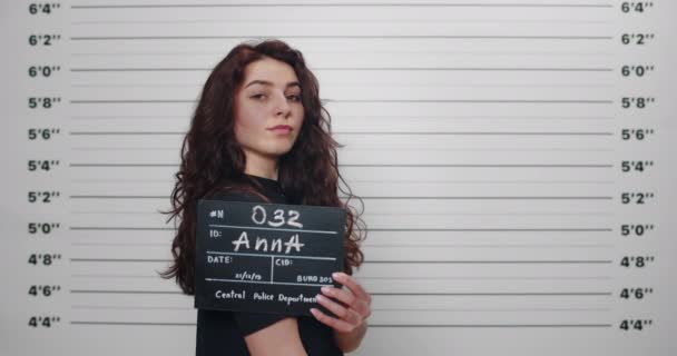 Boční profil přepadení mladé ženy s kudrnatými vlasy otáčející hlavu a dívá se do kamery. Žena zatčena s nápisemfotografována před policejní metrickou lineup stěnou. — Stock video