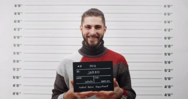 Polis merkezinde sakallı ve fotoğraf için tabela tutan yakışıklı erkek suçlu portresi. Kameraya bakarken poz veren ve gülümseyen genç adam görüntüsü..