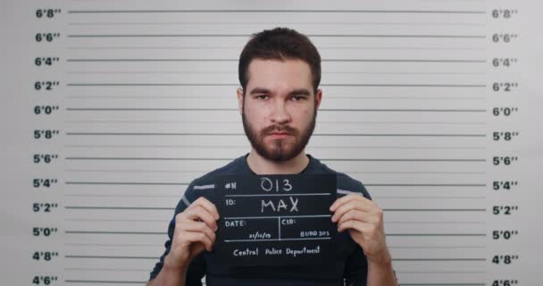 Portrait of young guy with mustaches and beard holding sign for photo in police department. Crop view of millennial man posing, raising head and looking to camera.Concept of criminal. — Stock Video