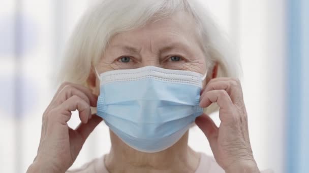 Крупный план зрелой пенсионерки с белыми волосами, снимающей медицинскую защитную маску и смотрящей в камеру. Портрет женщины-пенсионера, улыбающейся в больничной палате. — стоковое видео