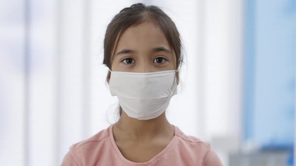 Retrato de niña en máscara protectora médica levantando la cabeza mientras mira a la cámara. Vista de cerca niño en el consultorio médico. — Vídeo de stock