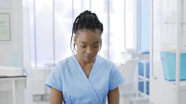 Vista de la cosecha de afro grave - médico americano mujer levantando la cabeza y mirando a la cámara. Retrato de mujer joven con rastas en uniforme médico en habitación de hospital. Concepto de asistencia sanitaria. — Vídeos de Stock