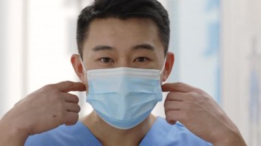 Asyalı doktorun tıbbi koruyucu maskeyi çıkartıp kameraya baktığı yakın çekim görüntüsü. Hastane odasında poz veren erkek doktor portresi. Sağlık hizmeti kavramı.