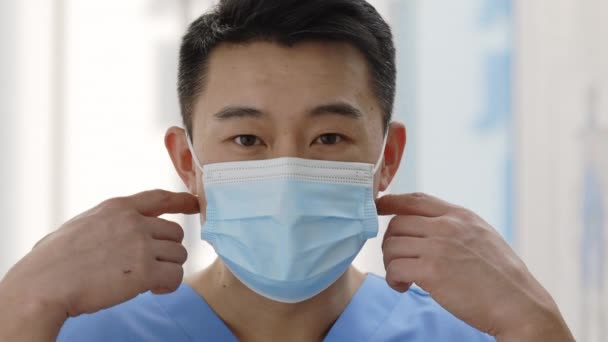 Közelről láthatjuk, ahogy az ázsiai orvos leveszi az orvosi védőmaszkot, miközben a kamerába néz. Egy férfi orvos portréja, aki a kórházi szobában pózol. Az egészségügyi ellátás fogalma. — Stock videók