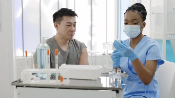 Schnittansicht einer Ärztin in Schutzmaske und Schutzhandschuhen, die eine Spritze mit Impfstoff füllt und mit einem männlichen Patienten im Krankenhauszimmer kommuniziert. Junger Mann wartet auf Impfung. — Stockvideo