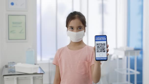 Ragazzina in maschera medica mostrando smartphone in mano con applicazione mobile per la vaccinazione. Vista di coltura di bambino femminile che tiene il passaporto digitale di persona vaccinata. Covid-19. — Video Stock