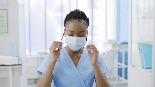 アフロの肖像-アメリカの女性看護師が医療保護マスクを脱ぐ。若いです女性のミステリービューでドレッドロックの頭を上げ、病院の部屋でカメラを見て. — ストック動画