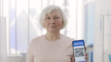 Yetişkin bir emekli kadın elinde akıllı telefon ve aşı için mobil uygulama taşıyor. Aşılanmış kişinin dijital pasaportuna sahip emekli bir kadının ekili görüntüsü.
