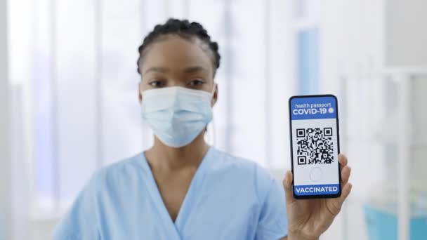 Giovane medico afro-americano in maschera medica che mostra smartphone in mano con applicazione mobile per la vaccinazione. Vista di coltura di infermiera femminile in possesso di passaporto digitale della persona vaccinata. Covid-19. — Video Stock