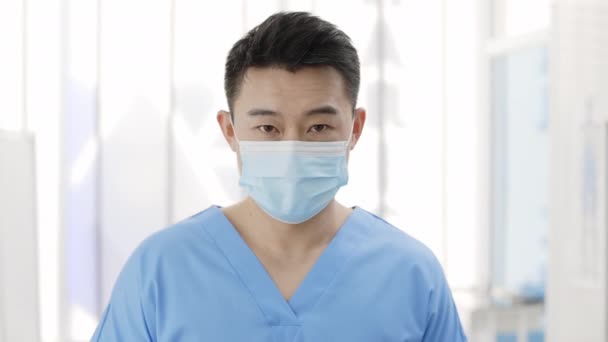 Tıbbi koruyucu maskeli Asyalı adamın kafasını kaldırıp kameraya bakarken görüntüsü. Hastane odasında poz veren erkek doktor portresi. Sağlık hizmeti kavramı. — Stok video