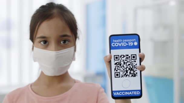 Ritratto di bambina in maschera medica che mostra smartphone in mano con applicazione mobile per la vaccinazione. Passaporto digitale immunitario per Covid-19. Codice QR. — Video Stock
