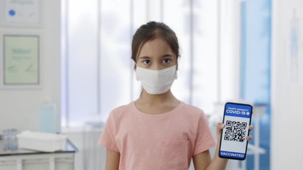 Immune passport. Young girl in medical mask raising up smartphone in her hand with mobile application for vaccination. Portrait of female child holding digital passport of vaccinated person. — Stock Video