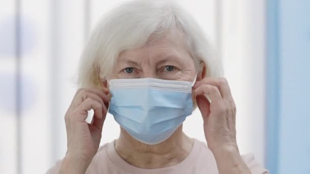 Beyaz saçlı yaşlı bir kadının tıbbi koruyucu maskesini çıkartıp kameraya bakarken görüntüsü. Hastane odasında emekli bir kadının portresi. Sağlık hizmeti kavramı, hasta. — Stok video