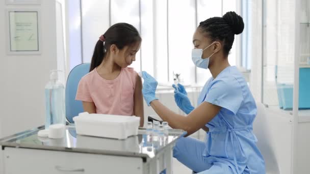 Vrouwelijke verpleegster met beschermend masker die tijdens het vaccinatieprogramma een injectie COVID-19 vaccin aan een klein meisje geeft. Begrip gezondheidszorg, preventie. — Stockvideo