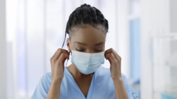 Közelkép a vegyes fajú női ápolónő bérbe fej és leveszi orvosi védő maszk, miközben nézi a kamera. Egy raszta hajú fiatal nő portréja az orvosi rendelőben. — Stock videók
