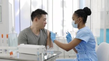 Asyalı hasta hastanede ya da klinikte iğne oluyor. Tıbbi maske takan Afrikalı-Amerikalı doktor kitle aşılama kampanyası sırasında genç bir adama grip ya da Covid-19 aşısı enjekte ediyor..