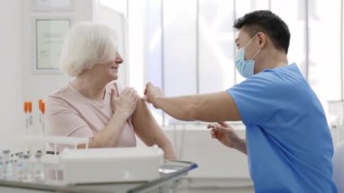 Koruyucu maskeli erkek doktor omzunu dezenfekte ederken ve aşı yaparken yaşlı bir kadının gülümsemesi. Enfeksiyon ve hastalık önleme kavramı.