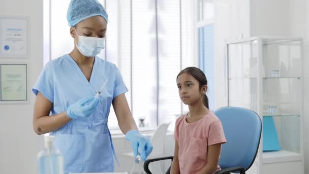 Koruyucu maskeli kadın hemşire, şapka ve güvenlik eldivenleri şırıngayı dolduruyor ve genç kız için aşı hazırlıyor. Çocuk hastanede sandalyede otururken aşı olmayı bekliyor.. — Stok video